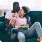Couple sits together on couch and shares a snack from a bowl.